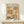 May include: Framed Paul Klee art print displayed on light wood shelf against cream wall. Abstract modernist artwork features geometric blocks in warm yellows, browns, and pinks with black line drawings of plants, figures, and architectural elements. Natural wood frame surrounds white matted print. Small white wrapped gift with greenery sits on shelf below artwork. Soft natural lighting creates gentle shadows on minimalist neutral setting.