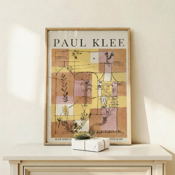 May include: Framed Paul Klee art print displayed on light wood shelf against cream wall. Abstract modernist artwork features geometric blocks in warm yellows, browns, and pinks with black line drawings of plants, figures, and architectural elements. Natural wood frame surrounds white matted print. Small white wrapped gift with greenery sits on shelf below artwork. Soft natural lighting creates gentle shadows on minimalist neutral setting.