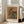 May include: Framed art print leaning against beige armchair in bright living room with white walls and windows. Print features Renoir's impressionist painting of pink and coral flowers in blue ceramic pitcher on warm golden background, with "VASE OF FLOWERS" and "PIERRE AUGUSTE RENOIR" text in clean modern typography above and below artwork.