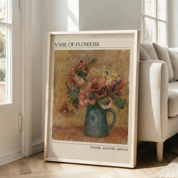 May include: Framed art print leaning against beige armchair in bright living room with white walls and windows. Print features Renoir's impressionist painting of pink and coral flowers in blue ceramic pitcher on warm golden background, with "VASE OF FLOWERS" and "PIERRE AUGUSTE RENOIR" text in clean modern typography above and below artwork.