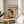May include: Framed art poster featuring Jackson Pollock's abstract expressionist drip painting with chaotic black lines, splatters and orange-red accents on cream background. Text reads "CHAOS AND CREATIVITY" and "POLLOCK" with dates "1912-1956". Thin wooden frame sits on round wooden side table next to beige upholstered chair with plant shadows casting across white wall in bright natural lighting.