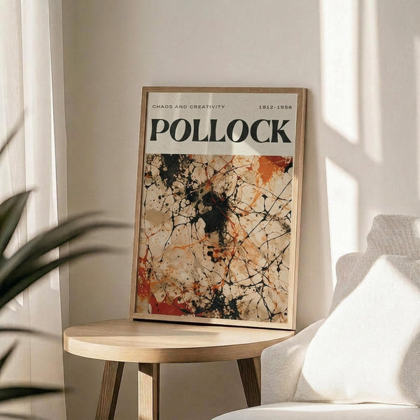 May include: Framed art poster featuring Jackson Pollock's abstract expressionist drip painting with chaotic black lines, splatters and orange-red accents on cream background. Text reads "CHAOS AND CREATIVITY" and "POLLOCK" with dates "1912-1956". Thin wooden frame sits on round wooden side table next to beige upholstered chair with plant shadows casting across white wall in bright natural lighting.