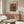 May include: A framed reproduction of Renoir's floral still life artwork showing colorful flowers in a blue ceramic pitcher, displayed above a mid-century modern walnut credenza with sliding doors. The warm-toned print features pink, orange and red blooms against a golden background. Scene includes a white upholstered chair, glass-top oval coffee table, and minimalist table lamp in a neutral living room setting.
