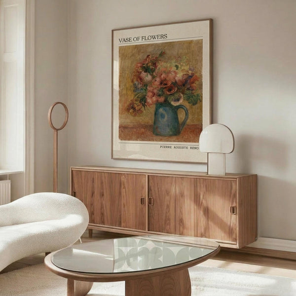 May include: A framed reproduction of Renoir's floral still life artwork showing colorful flowers in a blue ceramic pitcher, displayed above a mid-century modern walnut credenza with sliding doors. The warm-toned print features pink, orange and red blooms against a golden background. Scene includes a white upholstered chair, glass-top oval coffee table, and minimalist table lamp in a neutral living room setting.