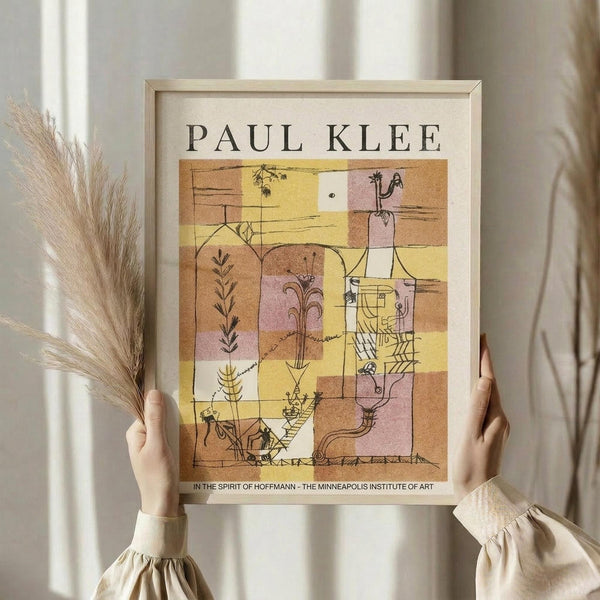 May include: Person in cream ruffled sleeves holding framed Paul Klee exhibition poster featuring abstract geometric composition in warm yellows, oranges, and pinks with botanical line drawings, alongside dried wheat stems against soft white curtained background