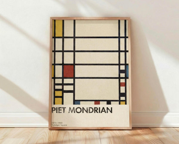 May include: Framed art print leaning against white textured wall on light wooden floor with soft natural lighting. The artwork features geometric abstract composition with black grid lines, cream background, and colored rectangles in yellow, red, and blue. Text below reads "PIET MONDRIAN" and "1872-1944 Trafalgar Square" in serif font. Natural wood frame visible around edges.