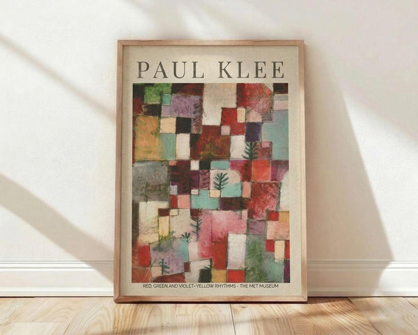 May include: Framed Paul Klee abstract art print featuring colorful geometric squares and rectangles in red, green, violet, yellow, and blue tones with small plant motifs scattered throughout. The poster has museum text at bottom and is displayed in a light wood frame against white walls on wooden floors with natural window lighting casting shadows.