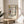 May include: Framed poster featuring Piet Mondrian's geometric abstract artwork with black grid lines and blocks of yellow, red, and blue on white background, displayed on a round wooden side table next to a beige upholstered chair with plant leaves visible in corner, all in a bright room with white walls and natural sunlight creating soft shadows.