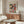 May include: Paul Klee abstract art poster with colorful geometric squares in reds, greens, and violets displayed above a wooden sideboard in modern living room. White upholstered chair and glass coffee table visible. Poster shows mosaic-like pattern with small evergreen tree motifs scattered throughout the squares. Natural lighting creates warm atmosphere with neutral wall colors.