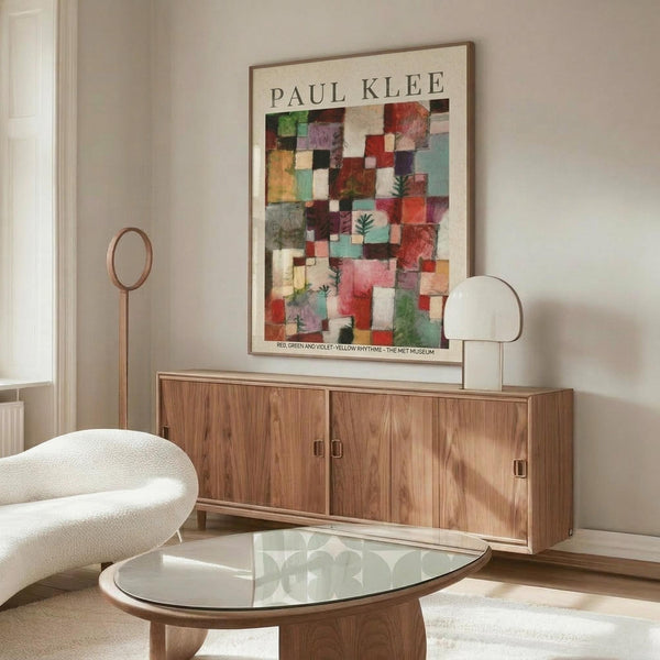 May include: Paul Klee abstract art poster with colorful geometric squares in reds, greens, and violets displayed above a wooden sideboard in modern living room. White upholstered chair and glass coffee table visible. Poster shows mosaic-like pattern with small evergreen tree motifs scattered throughout the squares. Natural lighting creates warm atmosphere with neutral wall colors.