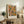 May include: Abstract expressionist art print in light wood frame displaying vibrant orange, blue, yellow and green paint splatters with black dots and lines. Poster sits on rustic wooden sideboard next to fern plant, ceramic vase, and stacked books in warm-toned living room setting with beige upholstered chair visible.