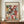 May include: A framed Paul Klee museum poster featuring an abstract composition of colorful geometric squares and rectangles in reds, greens, violets, blues, and earth tones with small evergreen tree motifs scattered throughout. The poster displays against cream walls with decorative molding, positioned on a wooden sideboard alongside a white ceramic vase and stacked books. The artwork shows varied textures and overlapping translucent layers creating depth and rhythm.