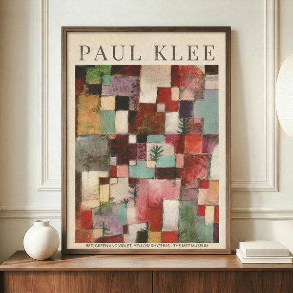May include: A framed Paul Klee museum poster featuring an abstract composition of colorful geometric squares and rectangles in reds, greens, violets, blues, and earth tones with small evergreen tree motifs scattered throughout. The poster displays against cream walls with decorative molding, positioned on a wooden sideboard alongside a white ceramic vase and stacked books. The artwork shows varied textures and overlapping translucent layers creating depth and rhythm.
