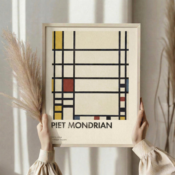 May include: Person in beige sweater holding framed Mondrian-style abstract art print featuring geometric grid pattern with black lines and colored rectangles in red, yellow, and blue on cream background. Light wood frame, pampas grass visible to left, soft natural lighting against white curtained window.