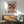 May include: Paul Klee abstract art poster with colorful geometric squares in reds, greens, violets and earth tones, featuring small botanical motifs. Framed print hangs above mid-century modern wooden credenza with white ceramic lamp. Sage green upholstered armchair with wooden frame sits beside round wooden side table. Bright natural light creates shadows on white wall in contemporary living room setting.
