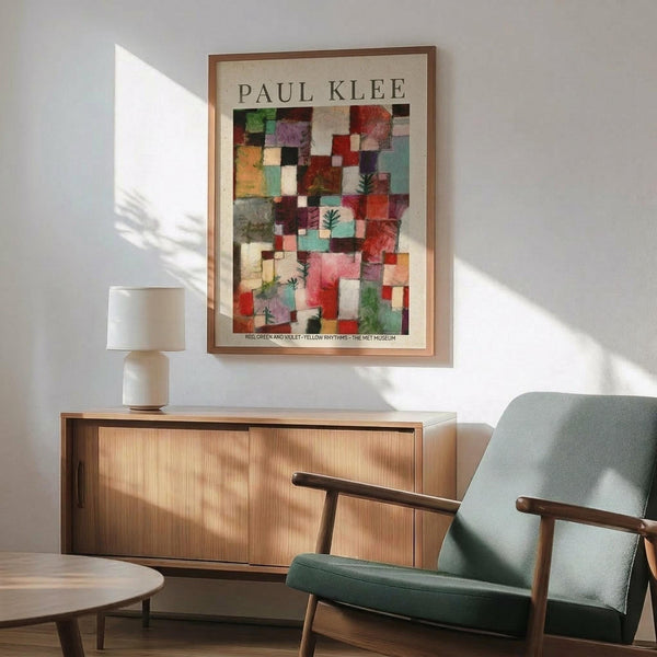 May include: Paul Klee abstract art poster with colorful geometric squares in reds, greens, violets and earth tones, featuring small botanical motifs. Framed print hangs above mid-century modern wooden credenza with white ceramic lamp. Sage green upholstered armchair with wooden frame sits beside round wooden side table. Bright natural light creates shadows on white wall in contemporary living room setting.