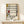 May include: Framed poster showing abstract geometric composition with black grid lines forming rectangles filled with white, yellow, red, and blue colors in Mondrian's distinctive style. Text below reads "PIET MONDRIAN 1872-1944 Trafalgar Square". Light wood frame sits on pale wooden surface against cream wall, with small white wrapped gift beside it featuring natural twine and dried sprig.