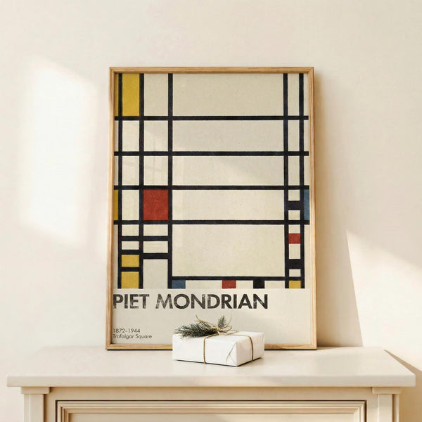 May include: Framed poster showing abstract geometric composition with black grid lines forming rectangles filled with white, yellow, red, and blue colors in Mondrian's distinctive style. Text below reads "PIET MONDRIAN 1872-1944 Trafalgar Square". Light wood frame sits on pale wooden surface against cream wall, with small white wrapped gift beside it featuring natural twine and dried sprig.