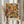 May include: Person holding framed abstract expressionist art print with vibrant orange, blue, yellow and green paint splatters and flowing black lines against cream background. Light wood frame, soft natural lighting, dried pampas grass visible to left, person wearing beige ruffled sleeve top.