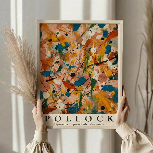 May include: Person holding framed abstract expressionist art print with vibrant orange, blue, yellow and green paint splatters and flowing black lines against cream background. Light wood frame, soft natural lighting, dried pampas grass visible to left, person wearing beige ruffled sleeve top.