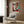 May include: Framed geometric abstract face artwork in Picasso-inspired cubist style with bold red, orange, teal and cream angular shapes forming fragmented facial features. White frame hangs on wood panel wall above beige upholstered armchair with geometric patterned pillow. Modern brass floor lamp and dark round side table with open book complete the contemporary living room setting with natural lighting.