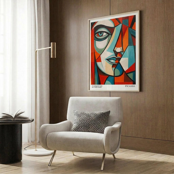 May include: Framed geometric abstract face artwork in Picasso-inspired cubist style with bold red, orange, teal and cream angular shapes forming fragmented facial features. White frame hangs on wood panel wall above beige upholstered armchair with geometric patterned pillow. Modern brass floor lamp and dark round side table with open book complete the contemporary living room setting with natural lighting.