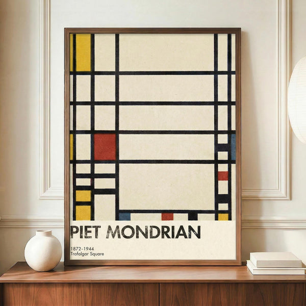 May include: Framed Mondrian abstract art print with geometric black grid lines and yellow, red, and blue color blocks on cream background, displayed on wooden sideboard next to white ceramic vase and stacked books in elegant room with white paneled walls