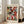 May include: A framed Paul Klee exhibition poster featuring an abstract composition of colorful geometric squares and rectangles in red, green, violet, pink, and turquoise with small evergreen tree motifs scattered throughout. The poster has "PAUL KLEE" at the top and "RED, GREEN AND VIOLET-YELLOW RHYTHMS - THE MET MUSEUM" at the bottom. Displayed in a light wood frame leaning against a white wall next to a beige upholstered sofa in a bright, modern living room.