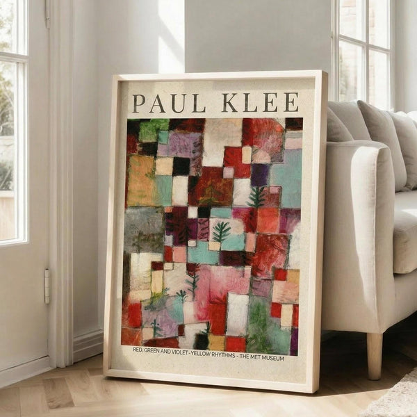 May include: A framed Paul Klee exhibition poster featuring an abstract composition of colorful geometric squares and rectangles in red, green, violet, pink, and turquoise with small evergreen tree motifs scattered throughout. The poster has "PAUL KLEE" at the top and "RED, GREEN AND VIOLET-YELLOW RHYTHMS - THE MET MUSEUM" at the bottom. Displayed in a light wood frame leaning against a white wall next to a beige upholstered sofa in a bright, modern living room.