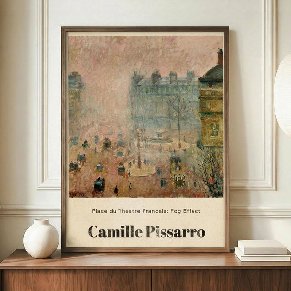 May include: Framed impressionist painting poster featuring a misty Parisian street scene with buildings, bare trees, and small figures walking through a foggy square. The artwork shows muted pink, blue and yellow tones with "Place du Theatre Francais: Fog Effect" and "Camille Pissarro" text below. Brown wooden frame sits on wooden sideboard next to white ceramic vase and stacked books against cream wall with white molding.