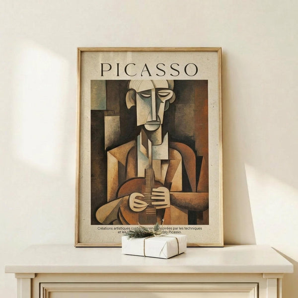 May include: Framed Picasso cubist artwork poster showing angular figure playing guitar in earth tones of brown, beige, and orange, displayed on light wooden console table next to small wrapped gift with greenery. Modern minimalist room setting with white walls and natural lighting creating soft shadows.