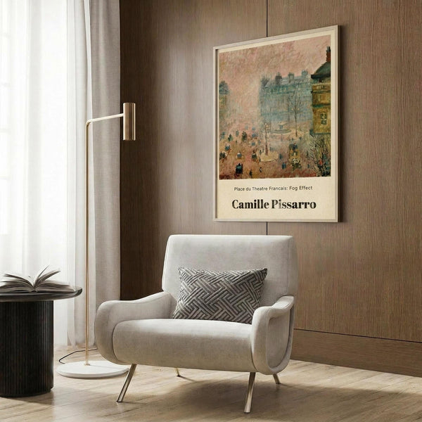 May include: Framed art print of Camille Pissarro's impressionist street scene with fog effect, showing Parisian buildings and figures in muted pink and blue tones. Print displayed on wood-paneled wall above beige upholstered armchair with geometric patterned pillow. Modern reading nook setting with brass floor lamp and side table.