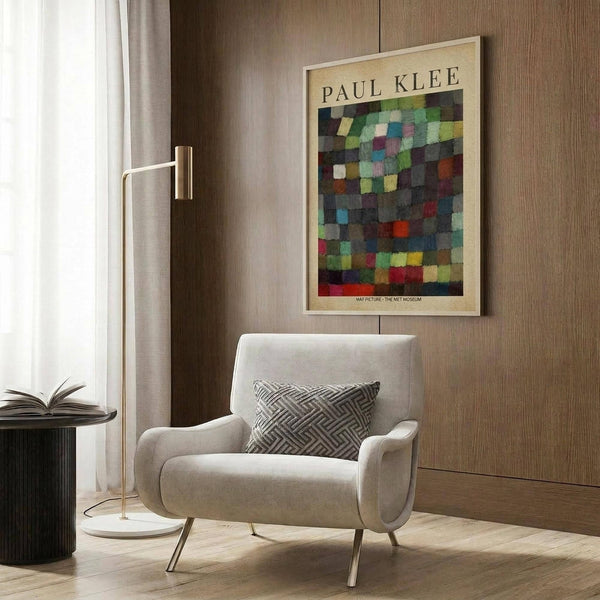 May include: Paul Klee abstract art print with colorful geometric squares in blues, greens, reds, and yellows, displayed in light wooden frame on wood-paneled wall. Modern reading nook features cream upholstered armchair with geometric patterned pillow, brass floor lamp, and dark side table with open book. Natural light from window with sheer curtains illuminates warm neutral interior space.
