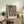 May include: Framed art print of woman in colorful traditional dress with red and orange striped skirt, displayed on rustic wooden console table alongside fern plant, ceramic vase, and books in neutral-toned living room setting with cream upholstered chair