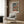 May include: Framed Picasso-style art poster showing woman in colorful traditional dress with red, blue, and orange tones, displayed above beige upholstered armchair with geometric patterned pillow. Modern living room setting features wood paneled wall, brass floor lamp, dark round side table with open book, and soft natural lighting from window with curtains.