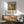 May include: Framed art print of impressionist street scene with misty fog effects, featuring people gathered in a plaza with historic buildings. Warm earth tones and muted blues. Displayed above modern wooden sideboard with white ceramic lamp, next to mid-century mint green armchair with wooden frame. Natural sunlight creates shadows across white wall in contemporary living room setting.
