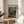 May include: Colorful abstract artwork poster featuring a grid of multicolored squares in blues, greens, reds, yellows, and earth tones, displayed in a light wood frame on a round wooden side table next to a beige upholstered chair. Soft natural lighting creates gentle shadows on white walls, with a plant visible in the corner.