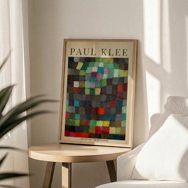 May include: Colorful abstract artwork poster featuring a grid of multicolored squares in blues, greens, reds, yellows, and earth tones, displayed in a light wood frame on a round wooden side table next to a beige upholstered chair. Soft natural lighting creates gentle shadows on white walls, with a plant visible in the corner.