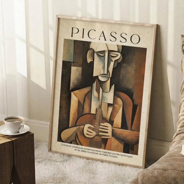 May include: Framed Picasso cubist artwork showing an abstract musician figure holding a guitar, rendered in earth tones of brown, beige, and muted colors. The poster leans against a white wall corner on a textured cream rug, with a coffee cup on a wooden block nearby and soft natural lighting creating shadows.