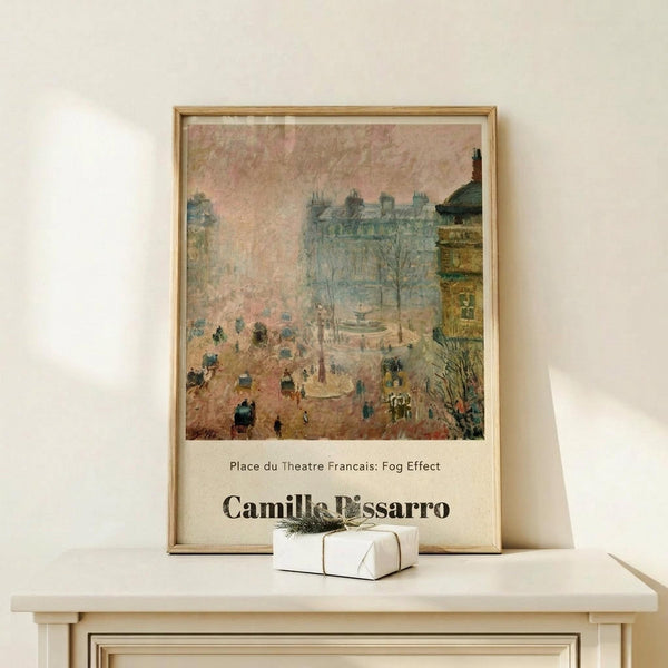 May include: Framed poster print displaying an impressionist painting of a foggy Parisian street scene with horse-drawn carriages, pedestrians, and classical architecture in muted pink and blue tones. The artwork shows Camille Pissarro's name at bottom. Light wooden frame sits on white wooden dresser next to small wrapped gift with twine and greenery sprig against cream wall.
