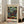 May include: Framed Paul Klee art print featuring colorful mosaic-like squares in muted greens, blues, purples, reds and earth tones, displayed in light wood frame against white wall next to beige sofa in bright room with large windows and polished floors.