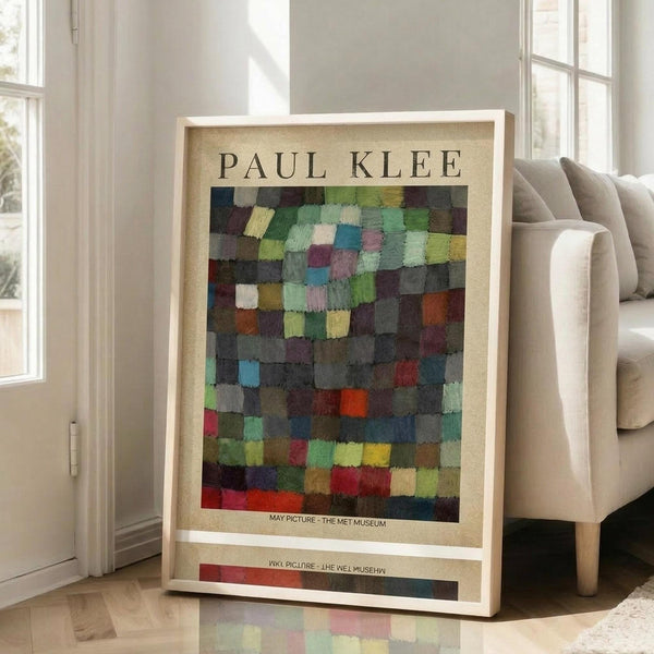 May include: Framed Paul Klee art print featuring colorful mosaic-like squares in muted greens, blues, purples, reds and earth tones, displayed in light wood frame against white wall next to beige sofa in bright room with large windows and polished floors.