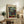 May include: Framed Paul Klee art print featuring colorful geometric squares in blues, greens, reds, and earth tones displayed on rustic wooden console with books, ceramic vase, and fern plant beside beige upholstered chair in neutral-toned living room setting.