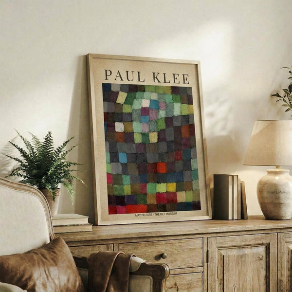 May include: Framed Paul Klee art print featuring colorful geometric squares in blues, greens, reds, and earth tones displayed on rustic wooden console with books, ceramic vase, and fern plant beside beige upholstered chair in neutral-toned living room setting.