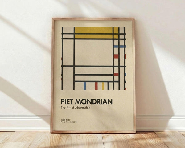 May include: Framed art poster leaning against white wall on light wood floor. Geometric abstract composition with black grid lines forming rectangles, filled with yellow, blue, and red color blocks on cream background. Text reads "PIET MONDRIAN The Art of Abstraction" with dates "1938-1943 Place de la Concorde" below. Natural wood frame, soft window light casting shadows across white textured wall.