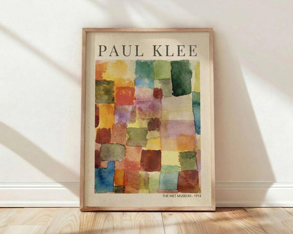 May include: Framed poster print featuring Paul Klee's abstract watercolor artwork with colorful geometric squares and rectangles in vibrant blues, greens, oranges, purples, and earth tones. The print shows "PAUL KLEE" at the top and "THE MET MUSEUM - 1914" at the bottom on cream-colored paper in a thin wooden frame, leaning against a white wall on light wooden flooring with natural sunlight casting diagonal shadows.