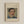 May include: Framed art print hanging on beige wall showing portrait of mustachioed man in blue jacket with yellow collar and white shirt. Cream textured mat displays painting in impressionist style with soft brushstrokes. Text above reads "Pierre Auguste Renoir painting" and below "portrait of a man". Light wood frame surrounds the piece in minimalist room setting.