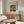 May include: Framed reproduction of Renoir portrait showing man with mustache in blue jacket, displayed above walnut credenza in minimalist living room with cream chair, glass coffee table, and white table lamp