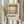 May include: Person in beige clothing holding framed abstract art poster featuring black grid lines with yellow, red, and blue geometric shapes on cream background. Light wood frame, pampas grass visible beside figure against neutral curtained backdrop.