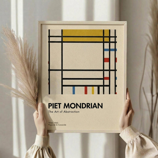 May include: Person in beige clothing holding framed abstract art poster featuring black grid lines with yellow, red, and blue geometric shapes on cream background. Light wood frame, pampas grass visible beside figure against neutral curtained backdrop.